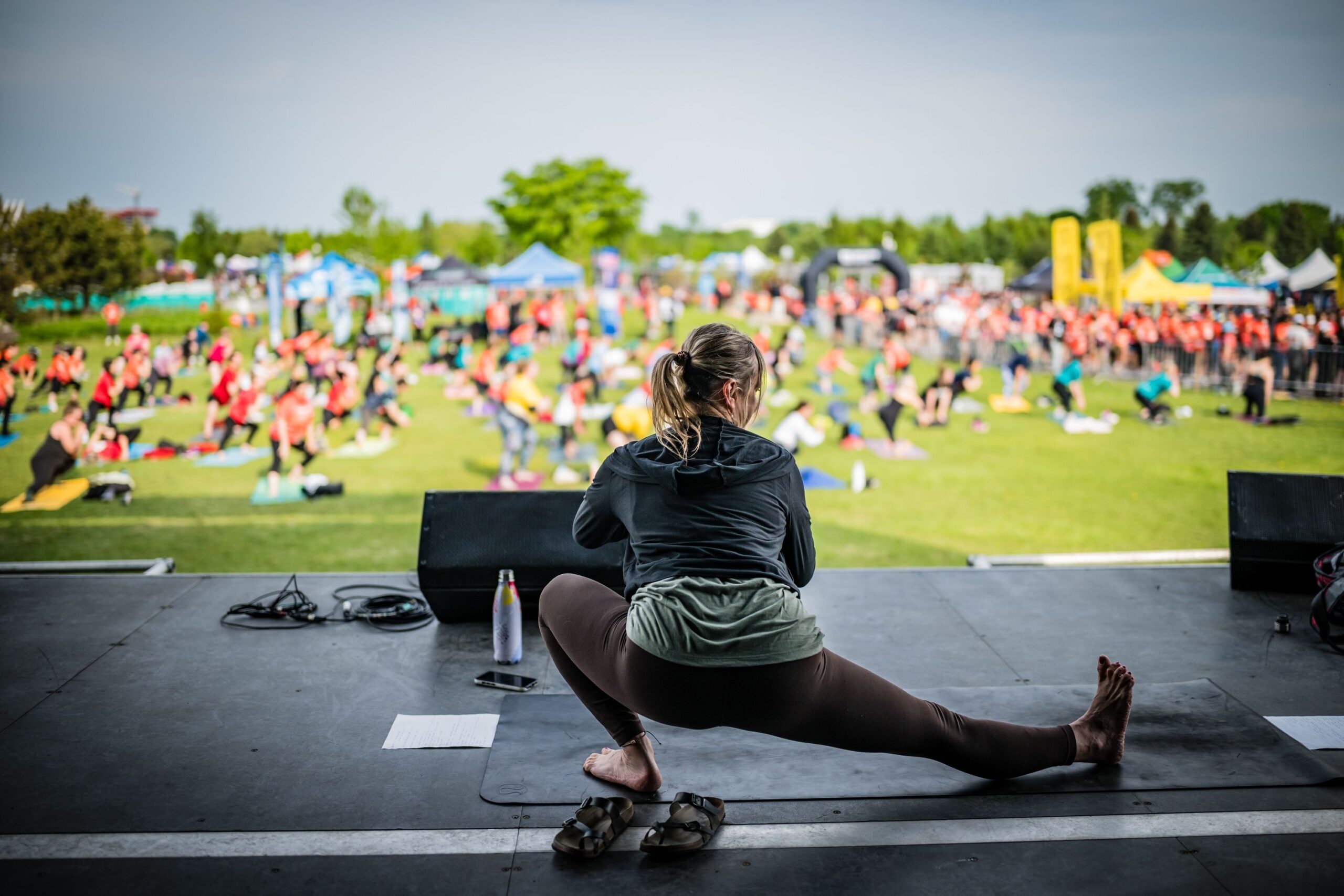 Person on stage in a wide-legged yoga stretch, watching a large crowd at an outdoor festival with tents in the background.